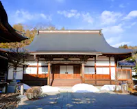 Traditional Japanese temple with a curved tiled roof, wooden pillars, and sliding doors, surrounded by small patches of snow and bare trees under a partly cloudy blue sky.