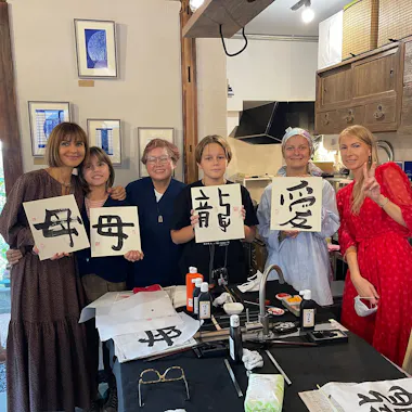 Calligraphy Workshop Five women stand together indoors, holding up paper sheets with Japanese calligraphy. Art supplies and more calligraphy papers are on the table in front of them. All are smiling, and the setting appears cozy and creative.
