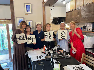 Five women stand together indoors, holding up paper sheets with Japanese calligraphy. Art supplies and more calligraphy papers are on the table in front of them. All are smiling, and the setting appears cozy and creative.
