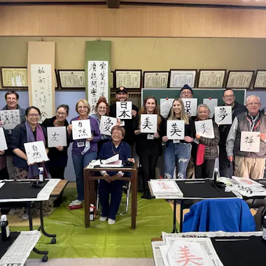 Calligraphy Workshop A group of people in a classroom hold up paper sheets with Japanese calligraphy. They smile at the camera, surrounded by brushes, ink, and completed calligraphy pieces displayed on the walls.