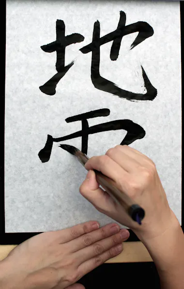 Calligraphy Workshop A person writes Chinese characters with a calligraphy brush on a sheet of white paper, using black ink. One hand holds the brush while the other steadies the paper.