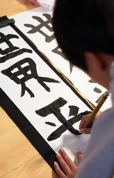 Calligraphy Workshop A child is practicing Japanese calligraphy on white paper with bold black characters, using a brush while kneeling on a wooden floor.