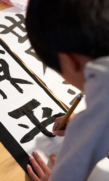 Calligraphy Workshop A child is practicing Japanese calligraphy on white paper with bold black characters, using a brush while kneeling on a wooden floor.