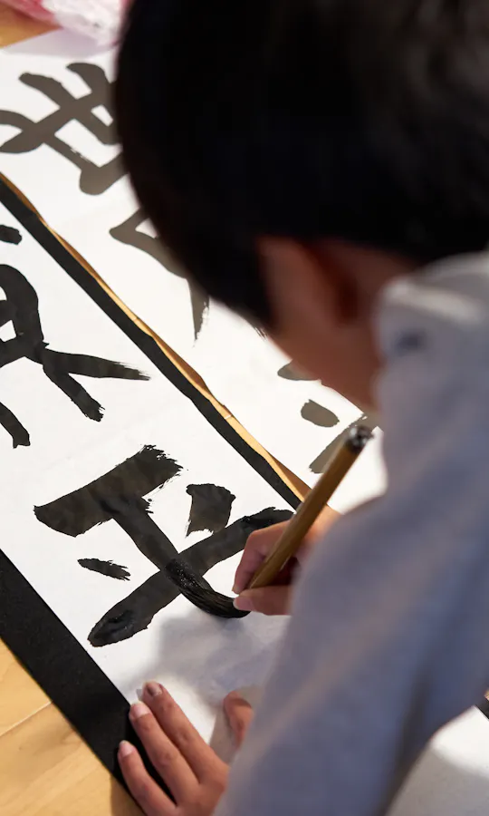 Calligraphy Workshop A child is practicing Japanese calligraphy on white paper with bold black characters, using a brush while kneeling on a wooden floor.