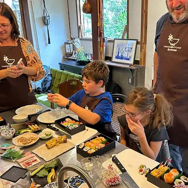 Cooking Workshop Two children and two adults gather around a kitchen table making sushi, with various ingredients, plates, and utensils spread out. The group appears focused and happy, enjoying the cooking activity together.