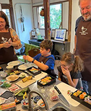 Two children and two adults gather around a kitchen table making sushi, with various ingredients, plates, and utensils spread out. The group appears focused and happy, enjoying the cooking activity together.