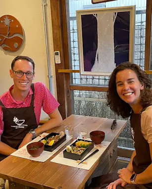Two people sit at a small wooden table in a cozy restaurant, smiling at the camera. They have bento boxes and soup bowls in front of them. A window and wall art are visible in the background.