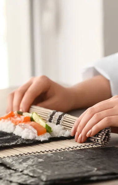 Making Sushi A person using a bamboo mat to roll sushi with seaweed, rice, and vegetables. Small bowls with ingredients are on the table beside them.
