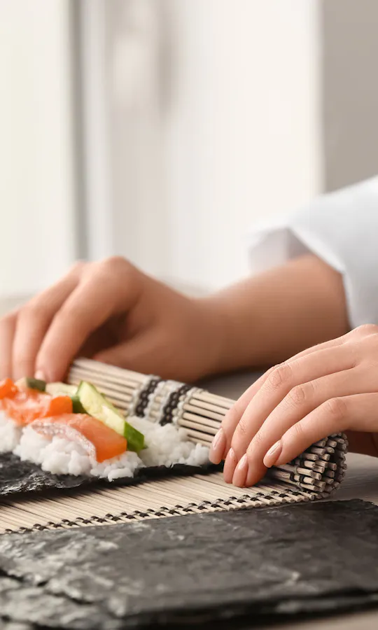 Making Sushi A person using a bamboo mat to roll sushi with seaweed, rice, and vegetables. Small bowls with ingredients are on the table beside them.