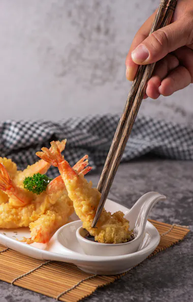 Tempura A hand uses chopsticks to dip a piece of shrimp tempura into sauce. More pieces of shrimp tempura are arranged on a white plate with garnish, set on a bamboo mat with a checkered cloth in the background.