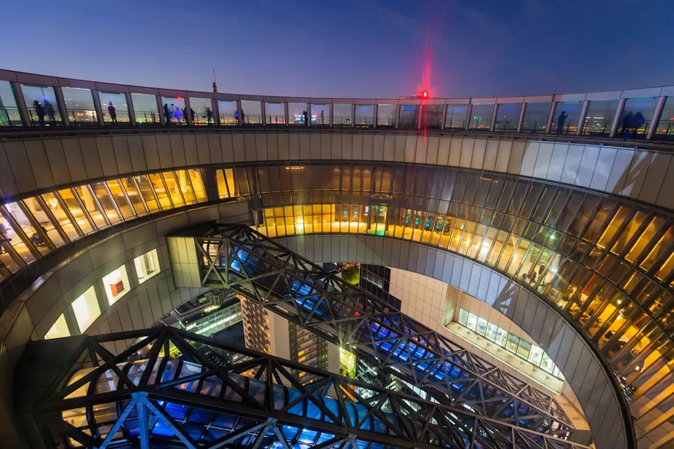 黄昏时分现代建筑结构的广角视图。圆形建筑采用多层设计，配有玻璃窗和照明内部。复杂的钢架结构和远处的天际线在黄昏的天空下增强了场景。