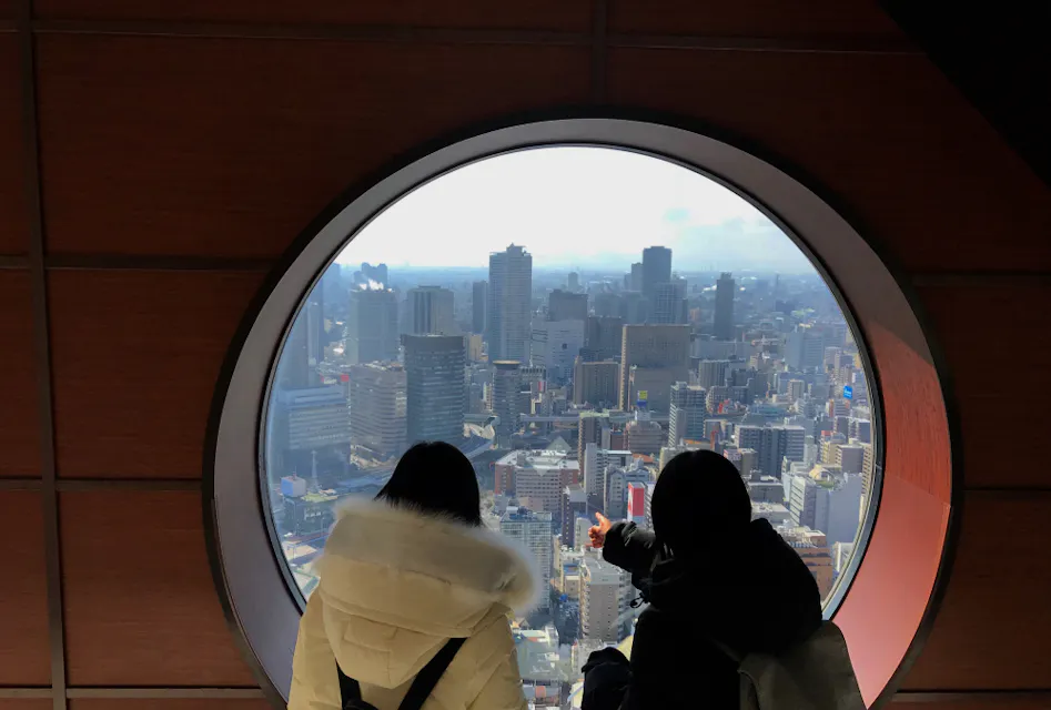 两个人穿着厚重的冬季外套，从后面看，透过一个大圆窗户向外望去，窗外是一个全景城市景观，包含众多建筑和部分多云的天空。场景被围绕窗户的室内墙壁框住。