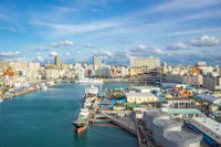 A harbor with boats docked beside industrial tanks and colorful containers, set against a cityscape of modern buildings under a bright, partly cloudy sky.