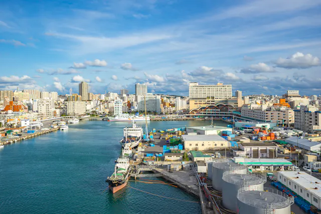 A harbor with boats docked beside industrial tanks and colorful containers, set against a cityscape of modern buildings under a bright, partly cloudy sky.