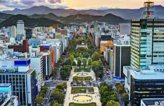 Aerial view of a green park with circular fountains and tree-lined paths, surrounded by tall buildings in a city, with mountains in the background at sunset.