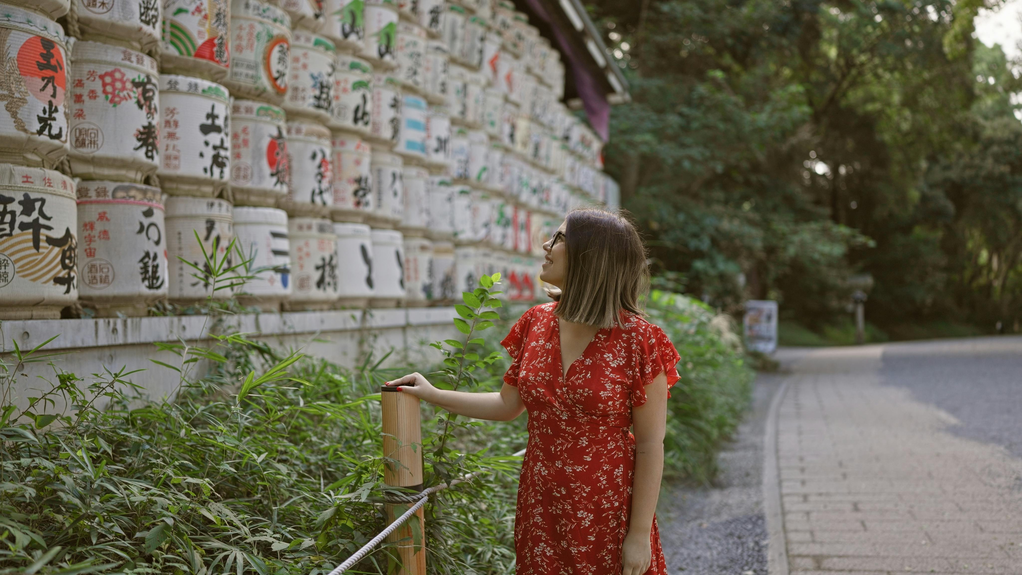 A woman in a red floral dress stands beside a wall of stacked decorative sake barrels with Japanese writing, surrounded by greenery on a paved path.