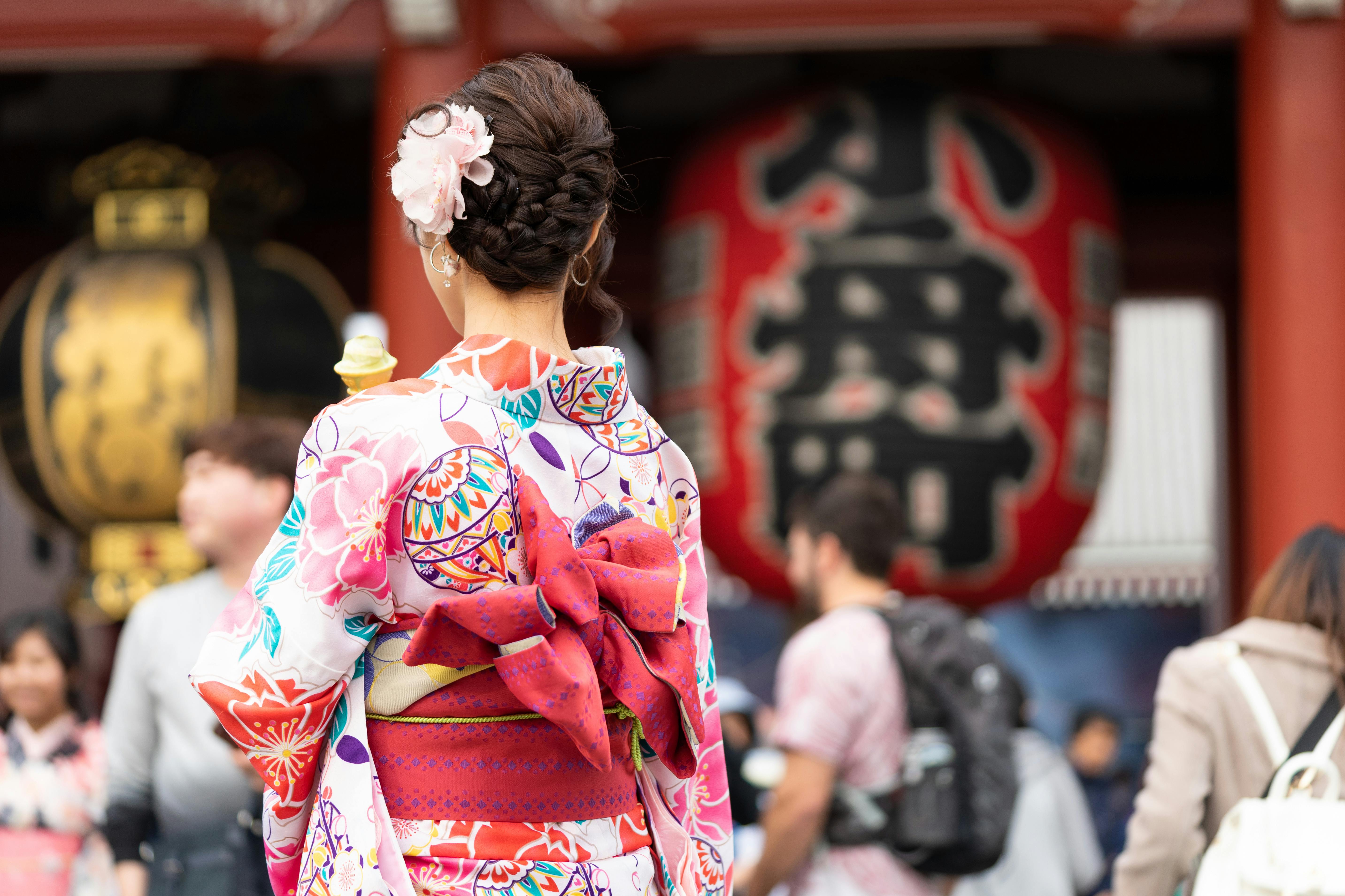 A woman wearing a colorful kimono with a large bow and a pink flower in her hair stands among a crowd near large traditional lanterns at a Japanese temple.