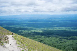 A winding mountain trail with two hikers leads down toward a vast, green forest under a mostly cloudy sky, with distant mountains visible on the horizon.