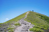 A narrow dirt trail leads up a grassy, sloped hill toward small structures under a clear blue sky. Sparse greenery surrounds the path.