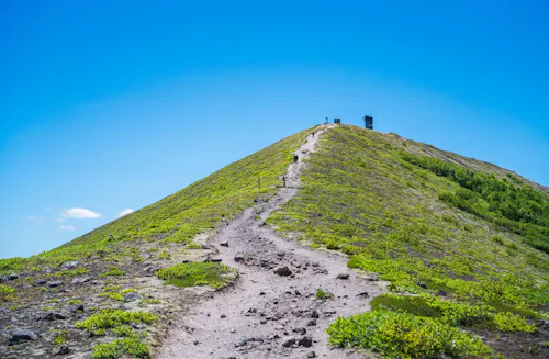 A narrow dirt trail leads up a grassy, sloped hill toward small structures under a clear blue sky. Sparse greenery surrounds the path.