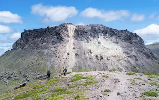 People hike toward a rocky, barren volcano summit under a bright blue sky with scattered clouds. Sparse green vegetation covers the foreground, contrasting with the dark volcanic rocks.