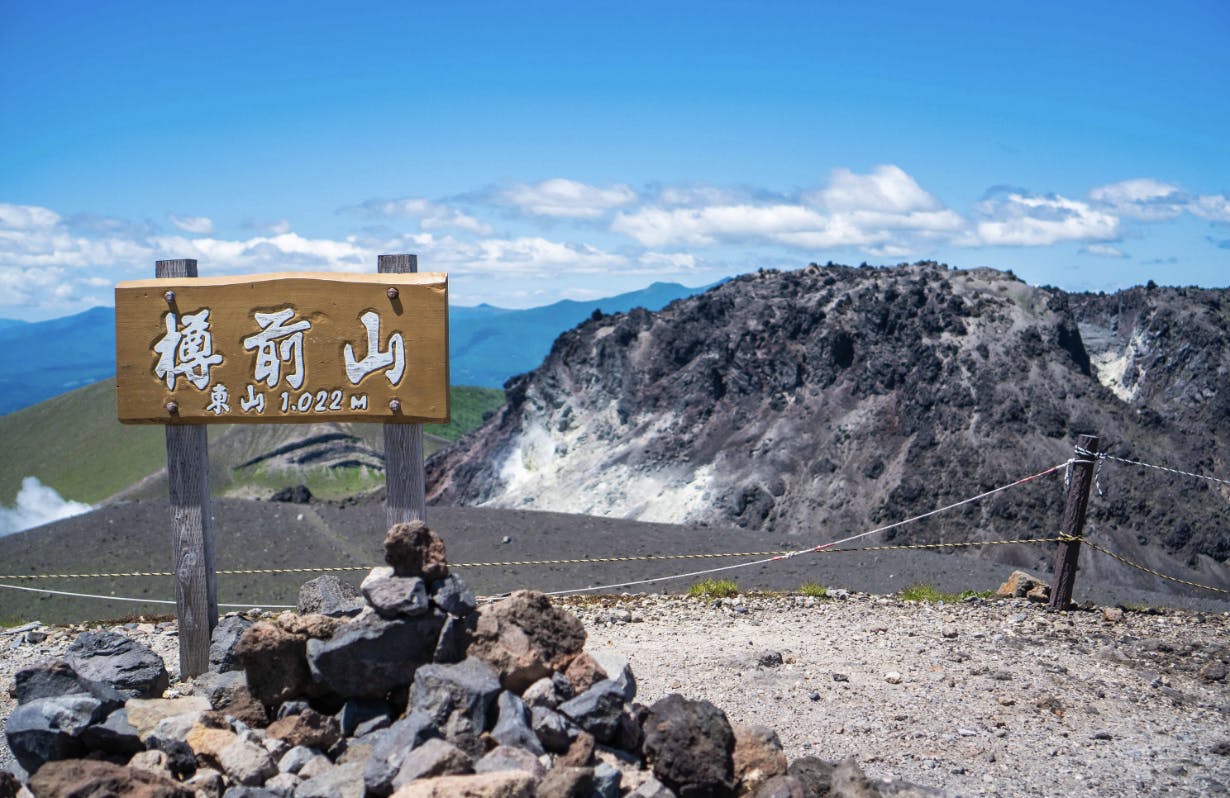 Wooden sign with Japanese characters and “1,022m” stands on rocky ground overlooking a mountainous volcanic landscape under a clear blue sky. Ropes line off the area near the sign.