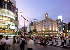 A busy city intersection at dusk with many people crossing the street, surrounded by illuminated buildings, shops, and a historic clock tower. The sky is purple and streetlights are lit.