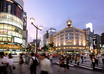 A busy city intersection at dusk with many people crossing the street, surrounded by illuminated buildings, shops, and a historic clock tower. The sky is purple and streetlights are lit.