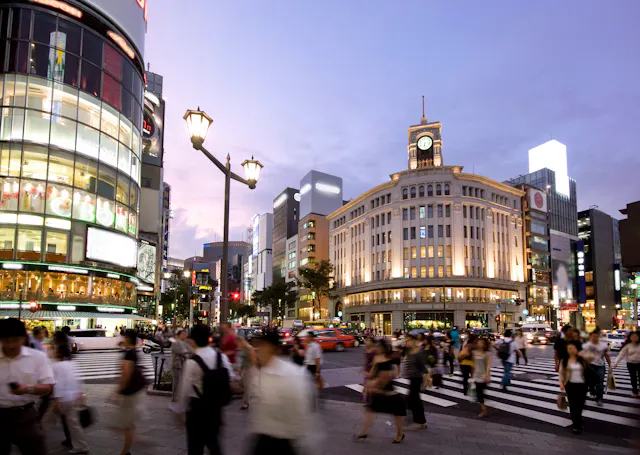 A busy city intersection at dusk with many people crossing the street, surrounded by illuminated buildings, shops, and a historic clock tower. The sky is purple and streetlights are lit.
