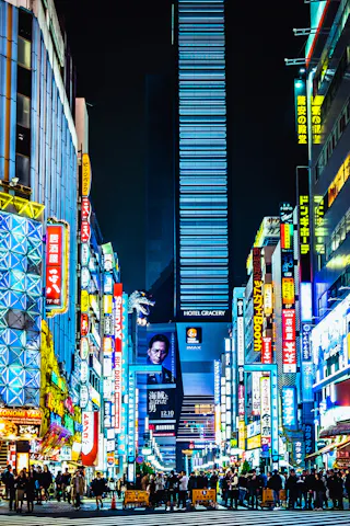 A busy street in Tokyo at night, lined with tall buildings covered in colorful neon signs and advertisements, crowded with people walking and gathering under the bright city lights.
