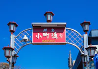 A red street sign reading "Komachi St." in English and Japanese, mounted on a decorative metal arch with lanterns, set against a clear blue sky.