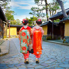 Nagamachi Two women in traditional Japanese kimonos walk side by side down a stone path, bordered by yellow walls and wooden buildings, with trees and a blue sky in the background.