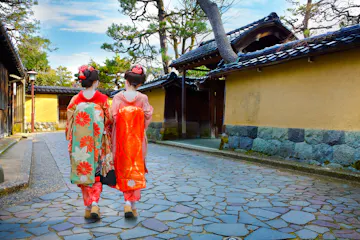 Nagamachi Two women in traditional Japanese kimonos walk side by side down a stone path, bordered by yellow walls and wooden buildings, with trees and a blue sky in the background.