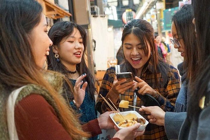 Four young women happily gather around a bowl of food with skewers at a market, with one taking a photo on her phone while the others smile and chat.