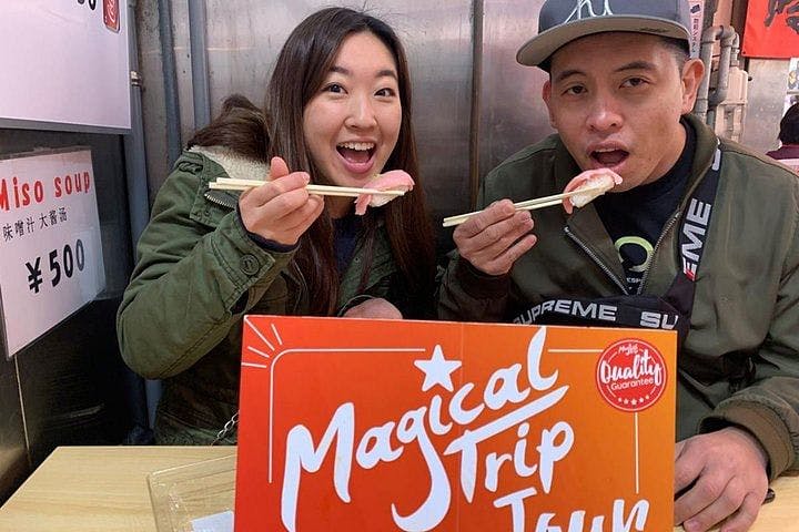 Two people sitting at a table, smiling and holding pieces of sushi with chopsticks. A red and white sign that reads "Magical Trip" is in front of them. A Miso soup sign with a price in yen is visible in the background.