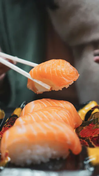 A close-up of salmon nigiri sushi, with chopsticks holding one piece above a tray containing more pieces of sushi. The focus is on the fresh, orange salmon slices atop white rice.