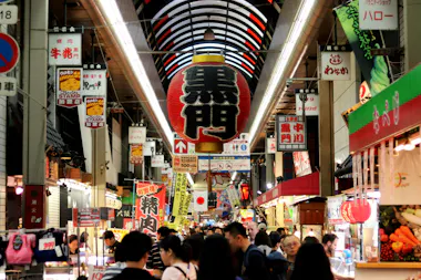 Crowd of people walking through a bustling indoor Japanese market lined with shops and colorful signs, including a large red lantern hanging from the ceiling with Japanese characters.