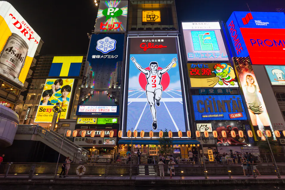 大阪の道頓堀での活気ある夜の風景。明るく照らされた広告やネオン看板が特徴。中央には、白と赤のランニングフィギュアが描かれたグリコのランニングマンの看板が目立つ。人々が通りの下で、賑やかな雰囲気を楽しんでいる。