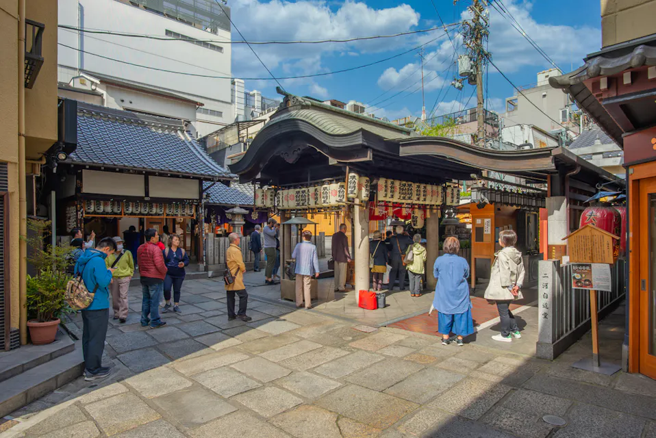 晴れた日に伝統的な日本の寺院を訪れる人々のグループ。祈っている人もいれば、寺院の中庭を歩いたり立ったりしている人もいます。建物は曲がった屋根と提灯を持つ古典的な日本の建築様式を示しています。