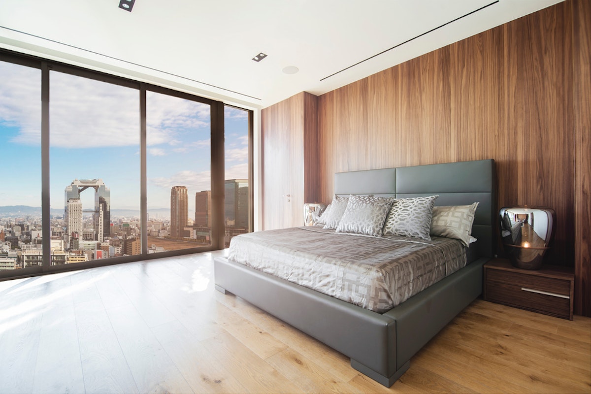 Interesting Osaka Hotels A modern bedroom with large floor-to-ceiling windows offering a panoramic city view. The room features a gray upholstered bed with patterned bedding, a wooden accent wall, and a sleek nightstand with a lamp. The floor is light wood. The overall ambiance is contemporary and chic.
