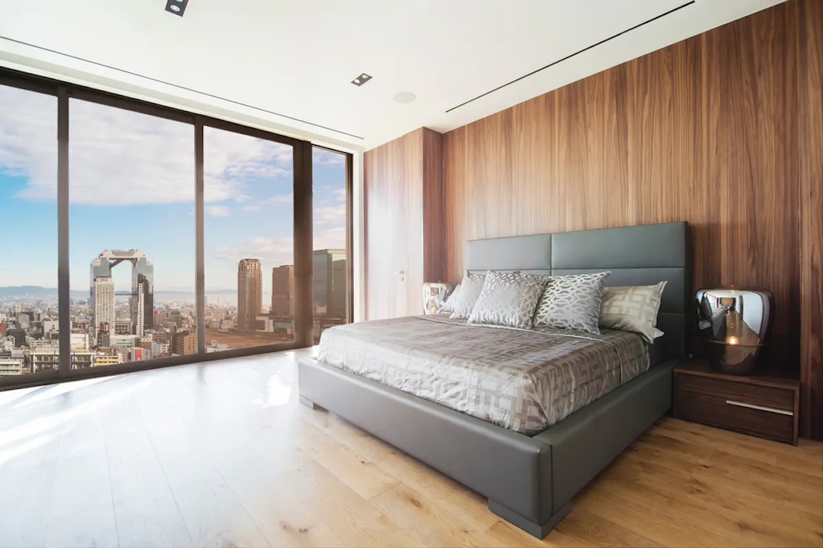 Interesting Osaka Hotels A modern bedroom with large floor-to-ceiling windows offering a panoramic city view. The room features a gray upholstered bed with patterned bedding, a wooden accent wall, and a sleek nightstand with a lamp. The floor is light wood. The overall ambiance is contemporary and chic.