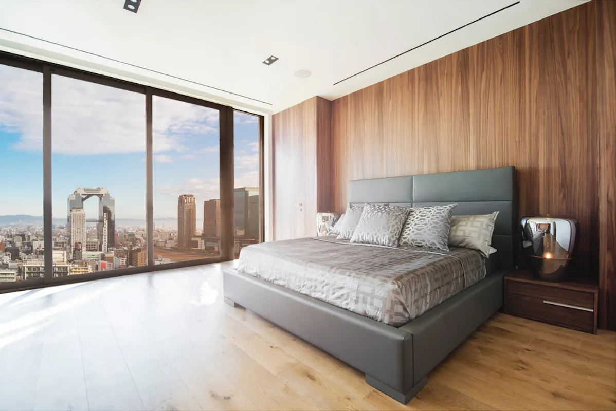 A modern bedroom with large floor-to-ceiling windows offering a panoramic city view. The room features a gray upholstered bed with patterned bedding, a wooden accent wall, and a sleek nightstand with a lamp. The floor is light wood. The overall ambiance is contemporary and chic.