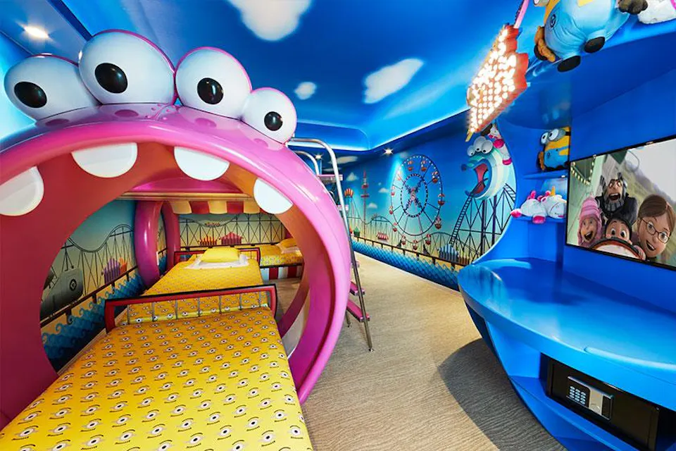 A vibrant, playful kids' bedroom with a colorful theme park mural. The room features a fun, monster-shaped bunk bed, with its mouth as the entrance. Bright blue walls and ceiling add cheerful vibes. A TV is mounted on the wall, displaying an animated film.