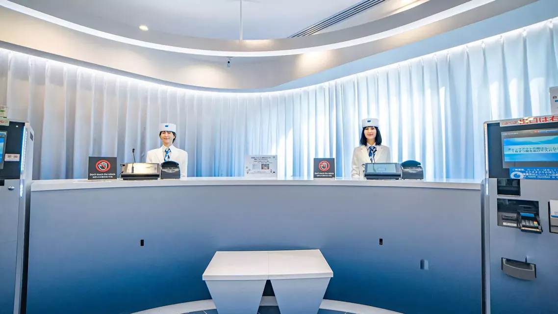 A futuristic hotel reception with two human-like robots in uniform standing behind a curved counter. The background features white, wavy curtains, and the counter has various signs and devices. A small triangular table is in front of the counter.