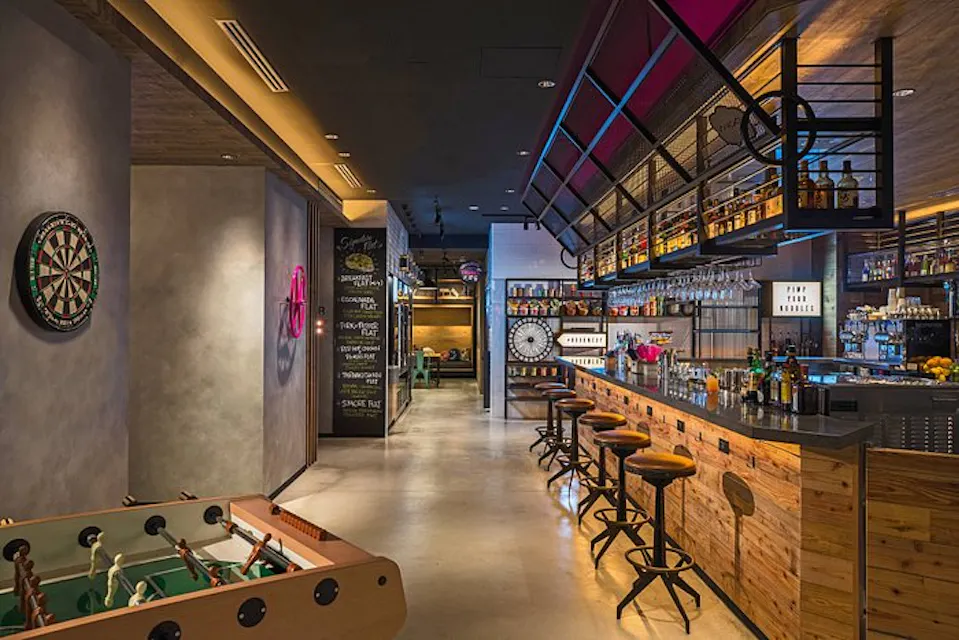 A modern bar interior featuring a long wooden counter with high stools, a wall-mounted dartboard, a foosball table in the foreground, and shelves filled with bottles above the bar. The space is warmly lit with various decorative items and a chalkboard in the background.