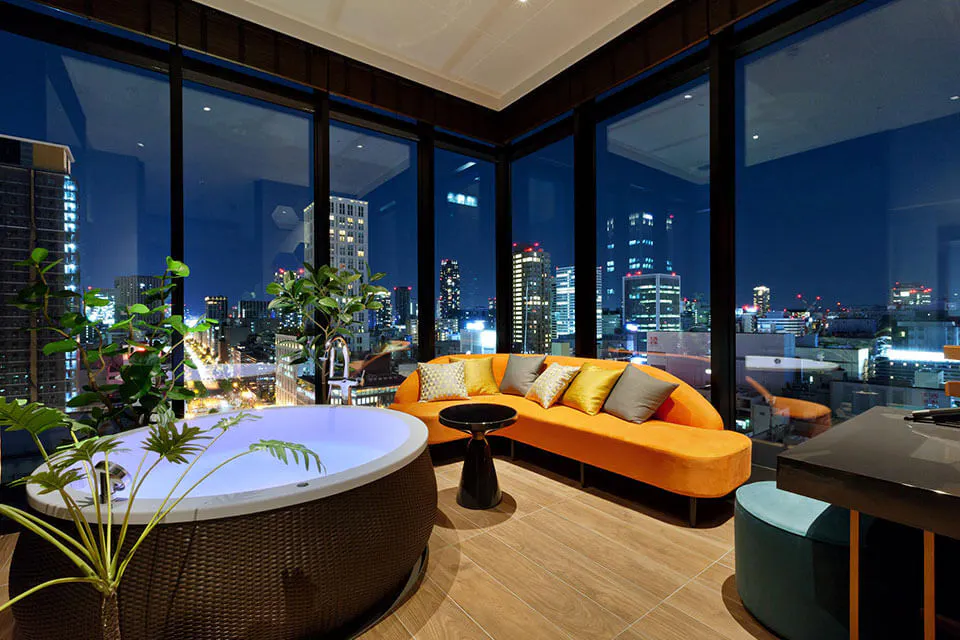 A luxurious living area with floor-to-ceiling windows offering a stunning cityscape view at night. The room features a circular soaking tub, an orange sectional couch with colorful pillows, a sleek black coffee table, and indoor plants, all under modern lighting.