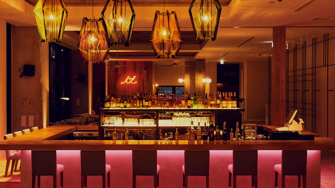 Modern bar interior with a long, illuminated counter lined with six bar stools. Shelves behind the counter showcase various bottles and glassware. Ambient lighting includes artistic hanging lamps, creating a cozy atmosphere. A neon sign in the background reads "it.