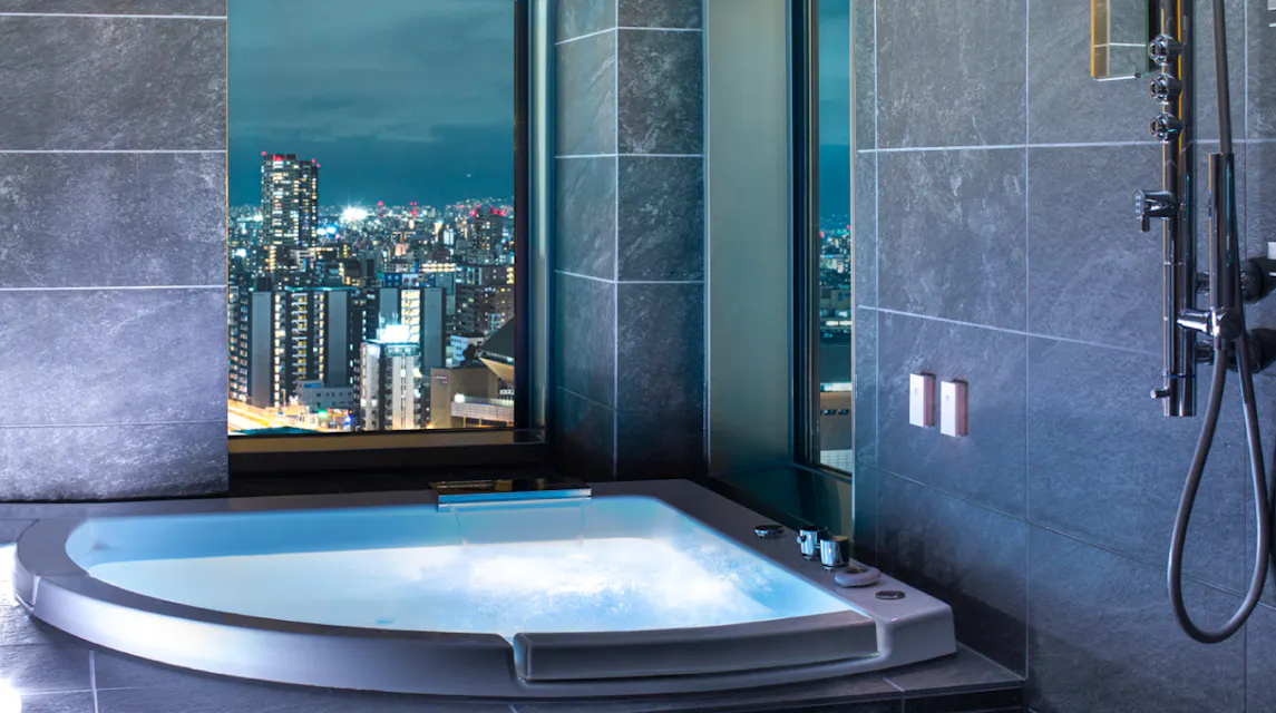A modern bathroom with a lit soaker tub, sleek dark tiles, and a large window offering a stunning night view of a city skyline with numerous illuminated buildings. The tub has a water faucet and various controls on the side.