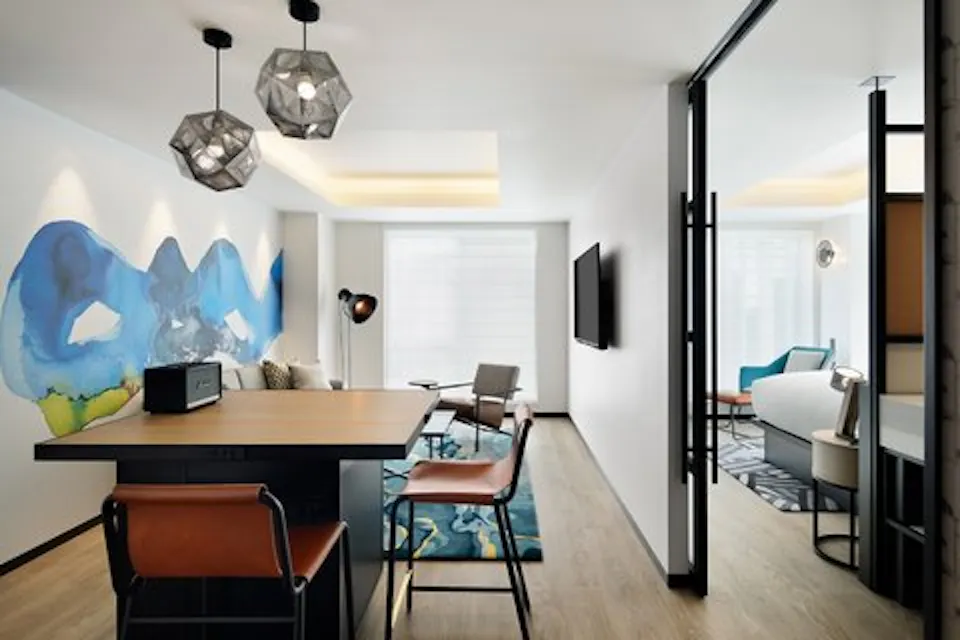 Modern apartment with an open layout. The living area features a geometric pendant light, abstract wall art, comfortable seating, and a TV. A sliding glass door leads to a brightly lit room with additional seating and large windows. The floors are light wood.