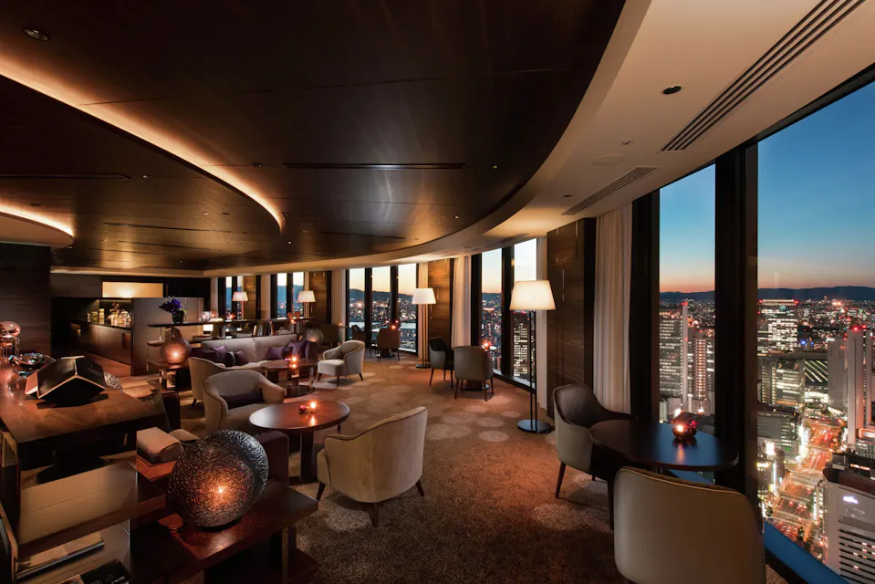Luxurious modern lounge with elegant furniture and ambient lighting, large floor-to-ceiling windows offering a panoramic view of a city's skyline at sunset.